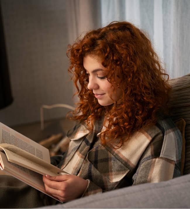 Person with curly red hair reading a book on a couch in softly lit indoor space