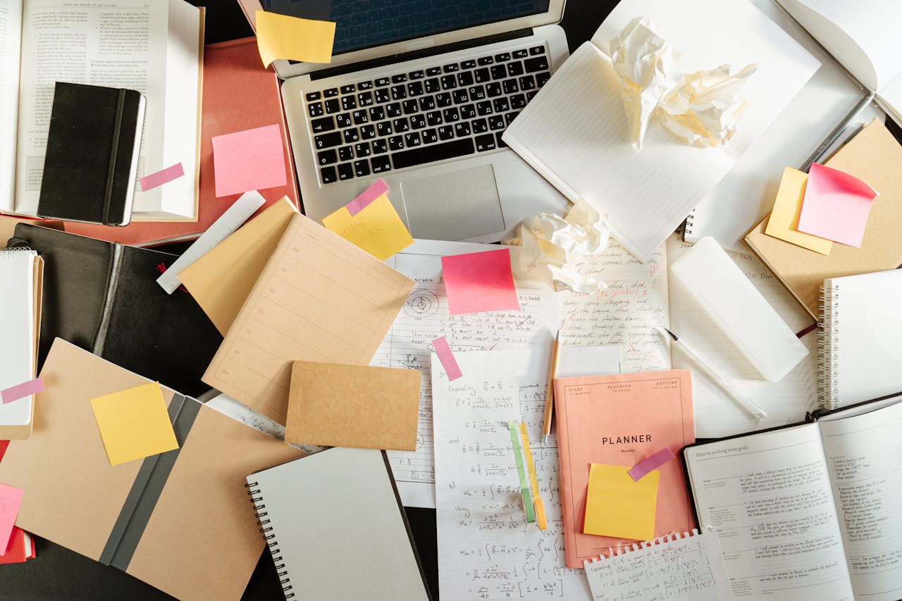 A cluttered desk with notebooks, planners, sticky notes, crumpled papers, and a laptop, suggesting a busy workspace or study area with scattered study materials and handwritten notes.