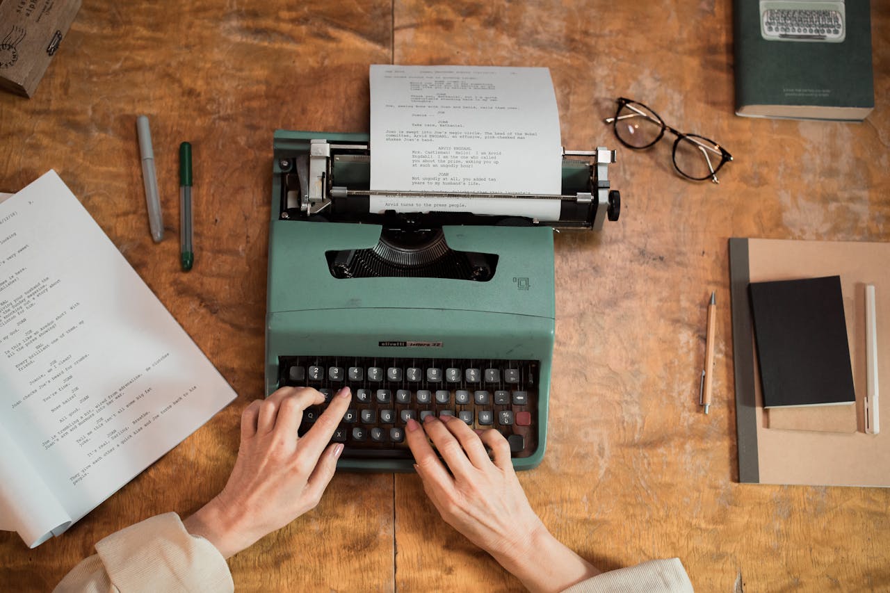 A person types on a vintage green typewriter with a script on it, surrounded by papers, pens, glasses, and a closed book on a wooden desk.