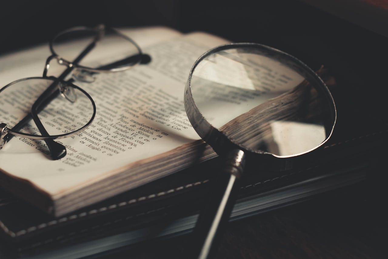 A pair of eyeglasses and a magnifying glass rest on an open book, with text clearly visible beneath the magnifying glass. The scene is softly lit, creating a warm, focused atmosphere for reading or research.