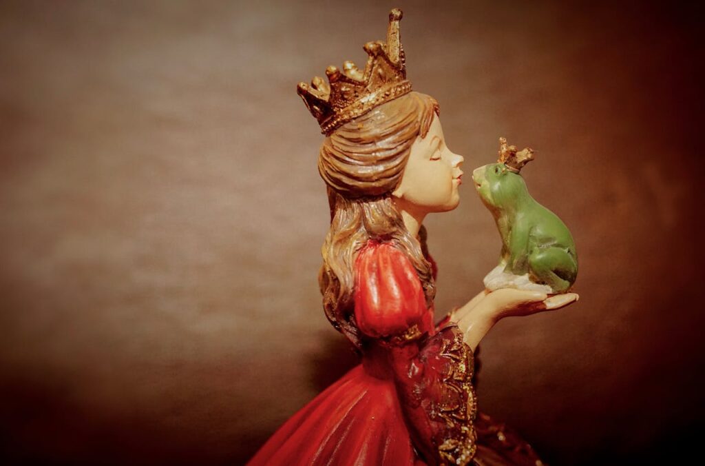 A statuette of a princess in a red gown with a gold crown, leaning in to kiss a small green frog also wearing a tiny crown, symbolizing a fairy tale or fantasy romance.