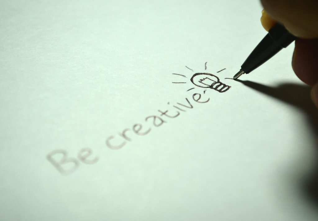 A hand holds a black pen, drawing a lightbulb icon next to the handwritten words 'Be creative' on a white sheet of paper, symbolizing the generation of ideas and effective use of writing prompts.