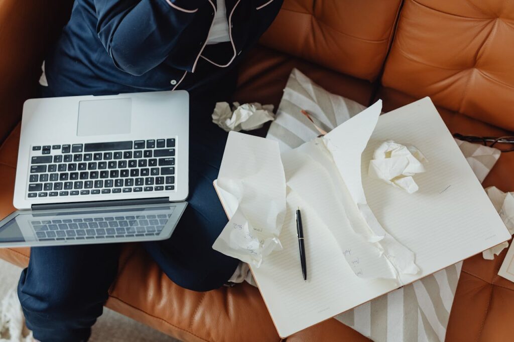 An overhead view of a person in pajamas sitting on a brown couch with a laptop on their lap, an open notebook with crumpled papers, and a pen beside them, ready to begin writing.