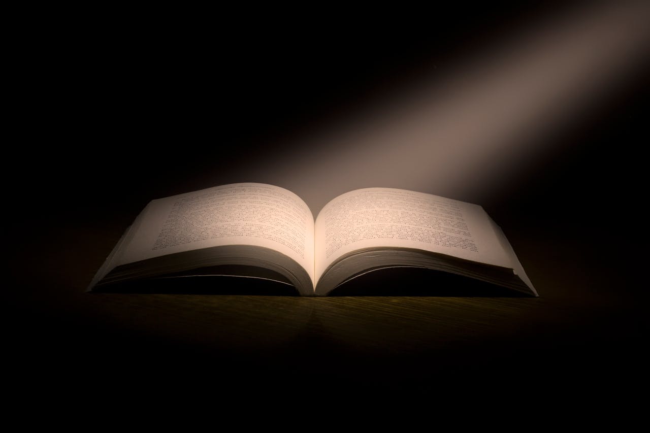 An open book with illuminated pages lies on a dark surface, dramatically lit by a single beam of light from above, symbolizing wisdom, hidden knowledge, or the power of stories.