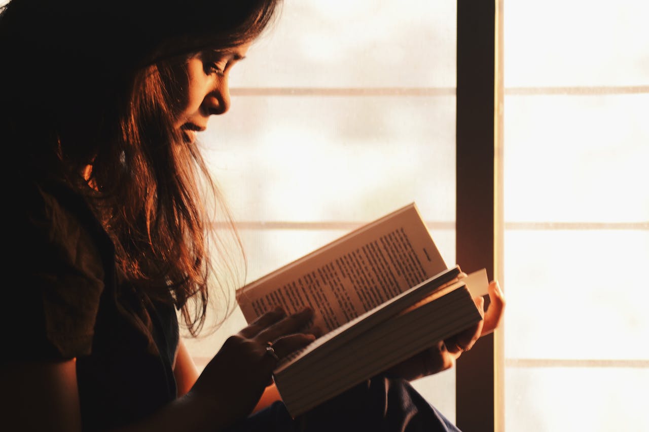 A woman with long hair sits by a sunlit window, deeply focused on reading a book. Warm light highlights her face and the pages she holds.