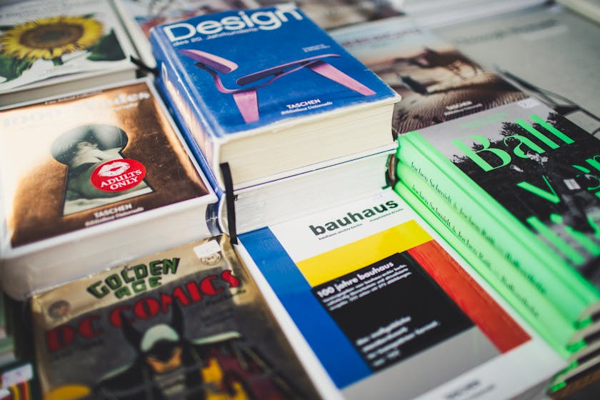 Flat lay of colorful books and magazines including Bauhaus, TASCHEN, and DC Comics, arranged in a grid pattern with bold cover designs.