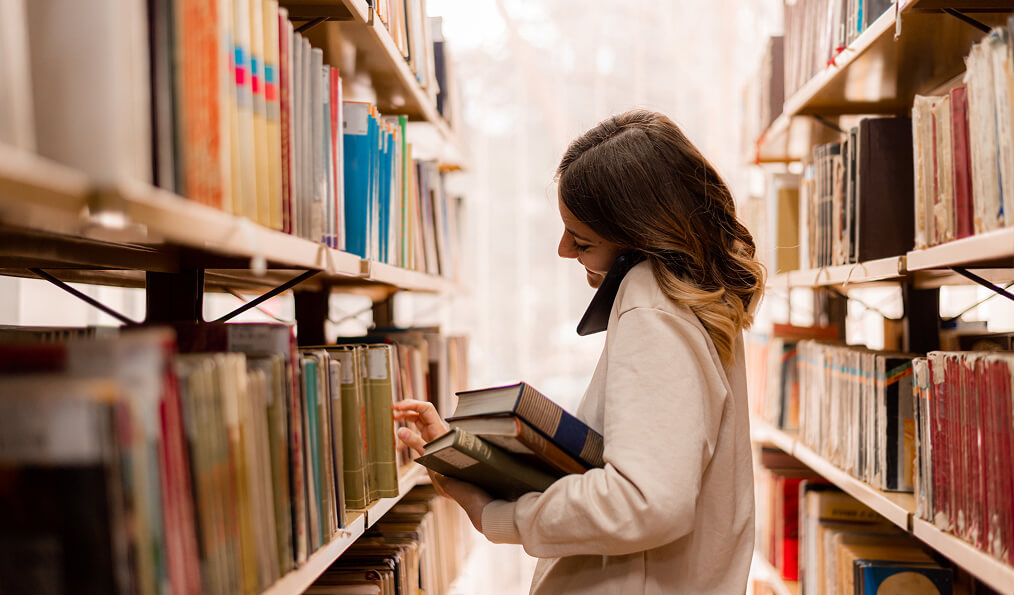A person stands between library shelves holding books and using a phone while reaching for another book.
