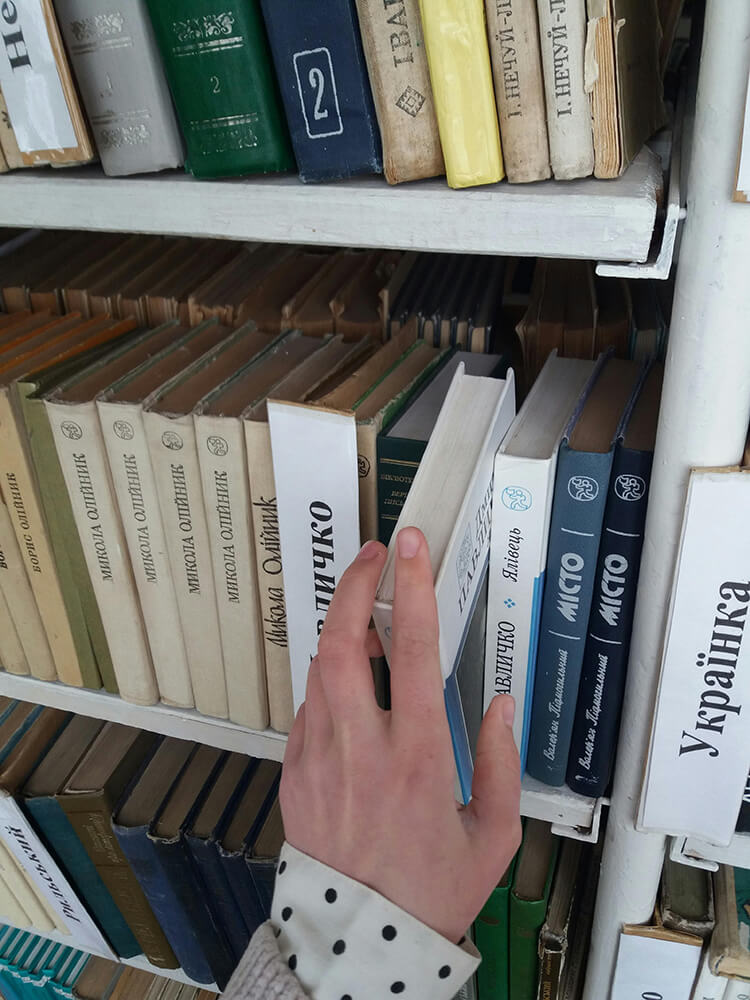 Close-up of a hand reaching for a book on a library shelf. The books are arranged vertically with Cyrillic titles, highlighting Ukrainian literature.