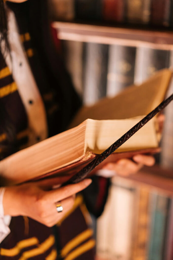 Person standing in front of a bookshelf, wearing a striped scarf and buttoned shirt, holding and reading a large thick book with a decorative cover.