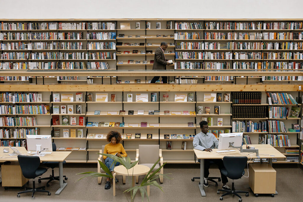 Interior of a modern library with multiple rows of bookshelves filled with books. One person browses the upper level, another sits reading in a chair, and a third works at a computer station.