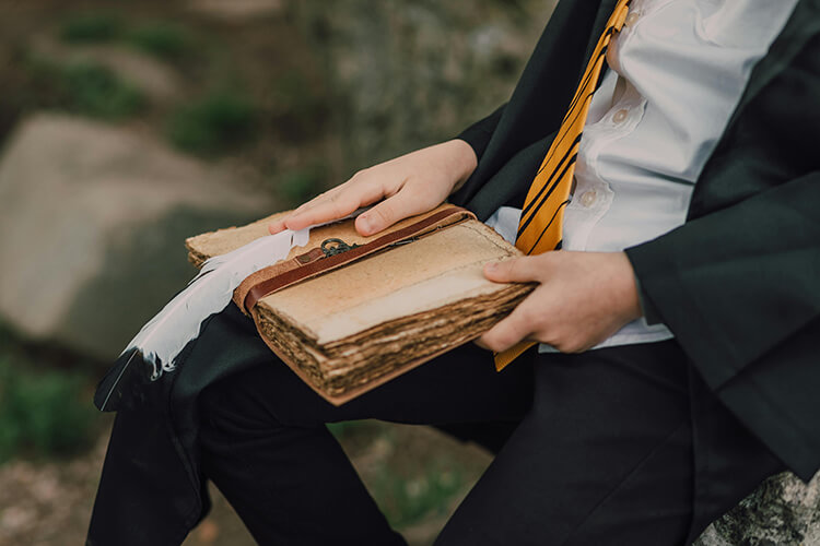 Person dressed in a white shirt, black robe, and yellow-and-black striped tie, holding an old weathered book with yellowed pages and a feather quill.
