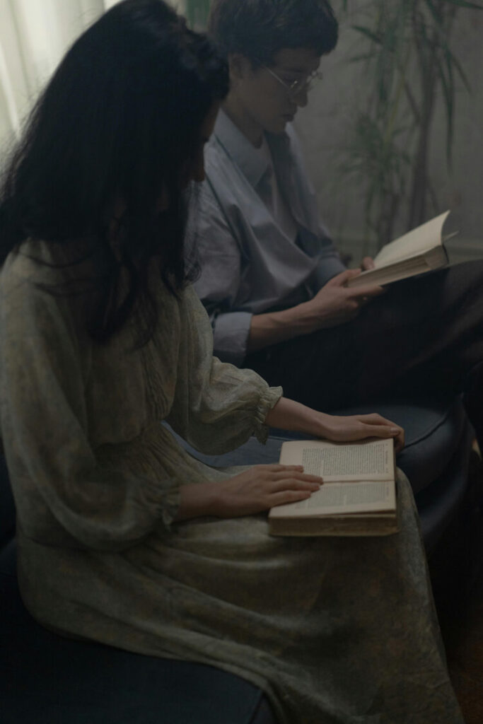 Two people sitting indoors, each reading a book. In the foreground, a person in a long light-colored dress with puffed sleeves sits on a dark chair holding an open book.