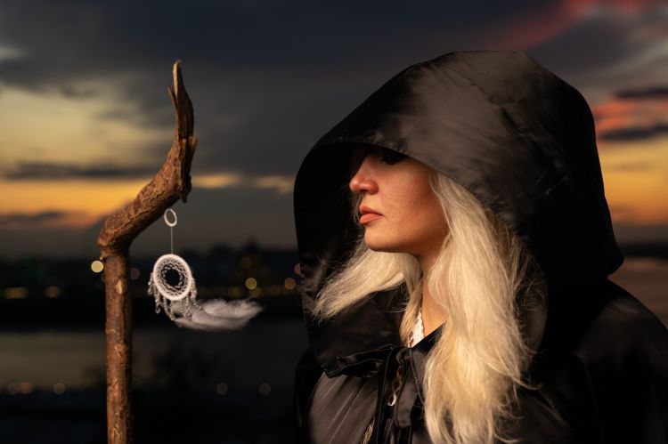 Woman in a hooded cloak holding a staff with sunset background.